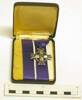 A New Zealand Memorial Cross medal with a thin mauve coloured ribbon attached. The medal is in the shape of a four pronged cross with a wreath design between the arms. A stem with seven leaves protrudes from the end of three arms of the cross. A crown sits atop one arm of the cross. The ribbon is wrapped around a felt covered backing. "GR VI" features at the centre. "21758 CPL A.M EAGLES" is engraved on the reverse of the medal. A cardboard rectangle is tucked behind the ribbon. "This Memorial Cross is forwarded to you/ by the Prime Minister on behalf of the/Government and people of New Zealand/ in memory of one who died in the service/of his country" is written on the cardboard. The medal is boxed inside a small black case with a hinged lid and a crown on the top.