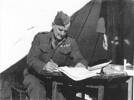 Portrait, WW2, Albert Henry Sage (12/3805 27881) seated at a wooden table, he has pencil in his hand and papers open on the table, he is outside a tent, his jacket has medal ribbons - This image may be subject to copyright