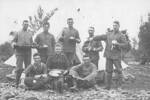 Group, soldiers with eating utensils, arranged for the photograph outside tent, foreground very rocky, seated soldier boots with hobnails, soldier pretending to tip liquid from cup onto another soldier seated, tin cups, plates, knife, metal roasting dish, Fred Baker standing back left holding metal roasting dish, other soldiers unknown. - No known copyright restrictions