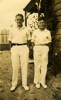 Jesse Pearce (left) a young boy standing wearing white shirt, long white trousers, white shoes [cricket whites] with a unidentified boy also in whites beside a wooden house - This image may be subject to copyright