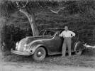 Collins, Tudor Washington (photographer). Portrait of Tudor Collins standing next to a Chrysler automobile. "X34.991" is the number plate of the vehicle. Rugs, or blankets, are on the grass in the background. Plants, a tree, a post and wire fence, a grass hill, are also in the background. Auckland War Memorial Museum - Tāmaki Paenga Hira. Tudor Collins Collection PH-2013-7-TC-B824-16.