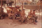 Members of 10 Pl relaxing at Kampong "DELTA".  R. T. Ormsby sits in front Kay Merito on guitar, J. Richardson seated on far right. A. McCutcheon in rear (second from left), Blackie Graham lying in hammock. M. Maytum directly behind Ormsby, David Neara clearning fun. Image taken during Malayan Emergency 1959-1960. © Peter Gallacher.